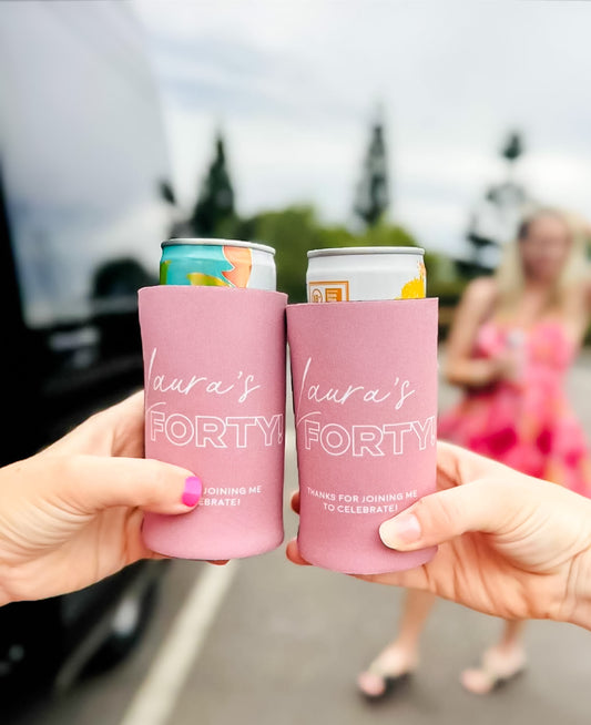 Personalised birthday stubby coolers. Two pink can coolers with 'Laura's Forty' text held by hands, blurred background of people and car. 