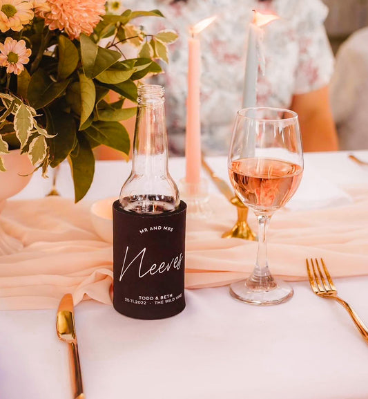personalised stubby cooler with 'Mr and Mrs Nevees' label on a wedding table setting with a glass of pink wine and floral arrangement.